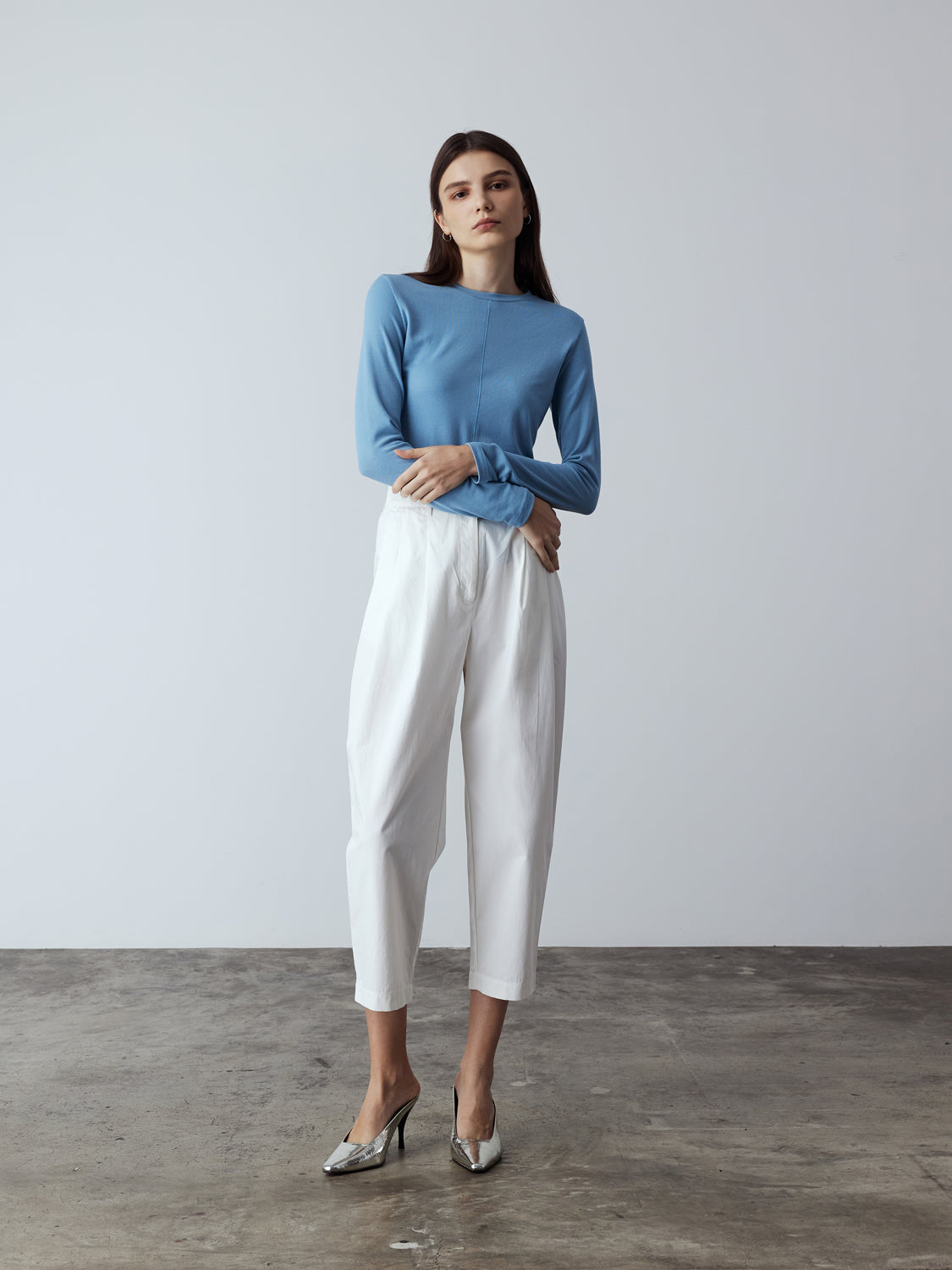 white : Model is wearing the Cotton Tailored Trousers in White, which comes high-waisted with slash pockets at the sides, a concealed zip-fly closure and pleated details. Worn with the Fitted Long Sleeve T-Shirt in Blue and metallic silver heels.