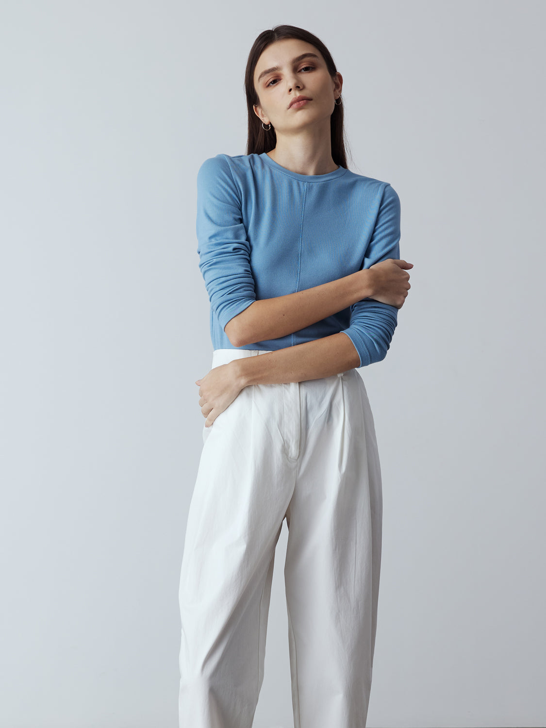 Model is wearing the Cotton Tailored Trousers in White, which comes high-waisted with slash pockets at the sides, a concealed zip-fly closure and pleated details. Worn with the Fitted Long Sleeve T-Shirt in Blue and metallic silver heels.