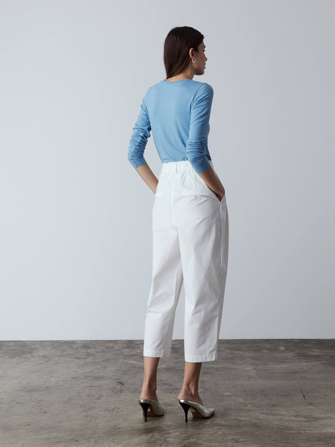 white : Model is wearing the Cotton Tailored Trousers in White, which comes high-waisted with slash pockets at the sides, a concealed zip-fly closure and pleated details. Worn with the Fitted Long Sleeve T-Shirt in Blue and metallic silver heels.