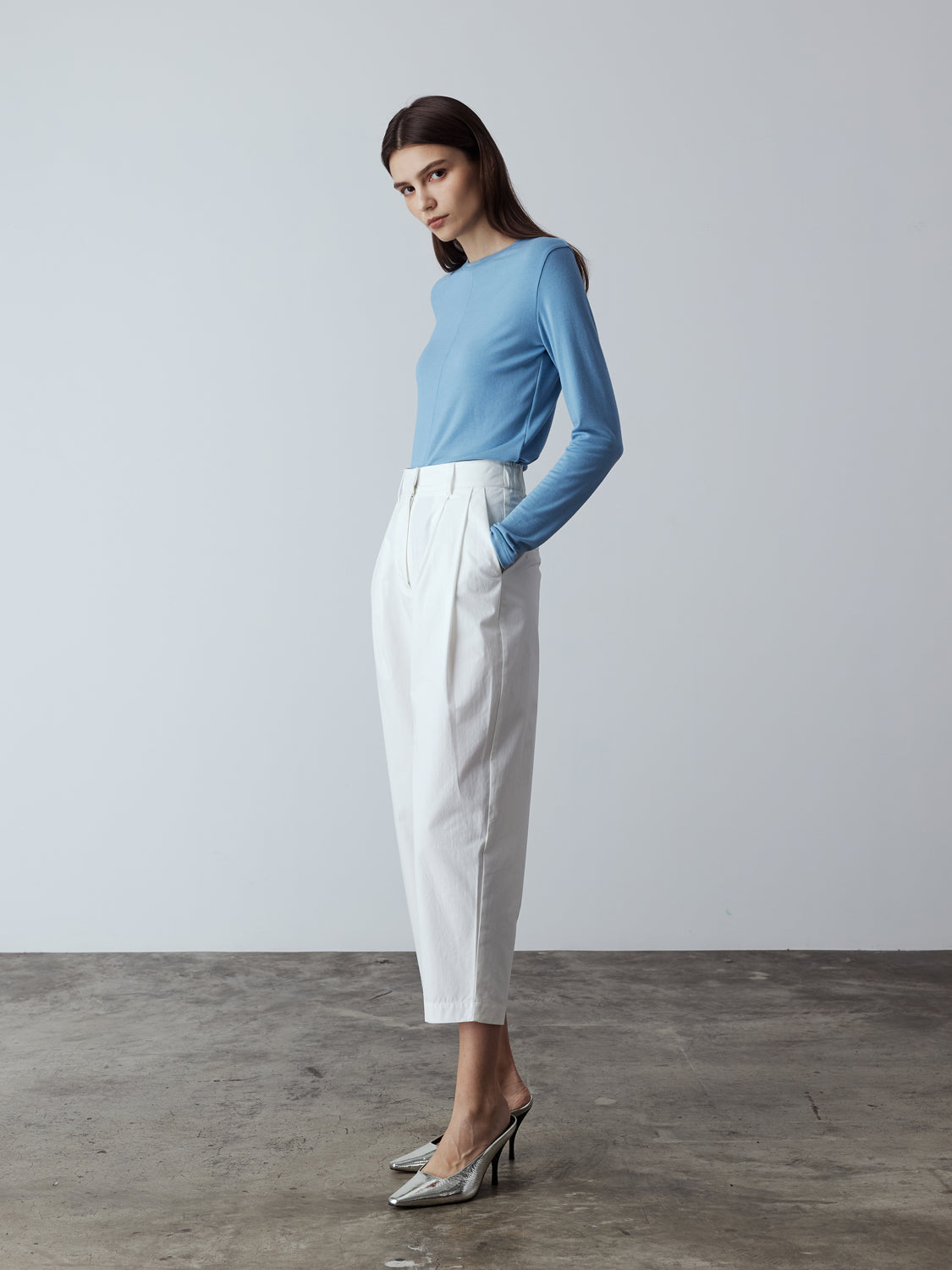 Model is wearing the Cotton Tailored Trousers in White, which comes high-waisted with slash pockets at the sides, a concealed zip-fly closure and pleated details. Worn with the Fitted Long Sleeve T-Shirt in Blue and metallic silver heels.
